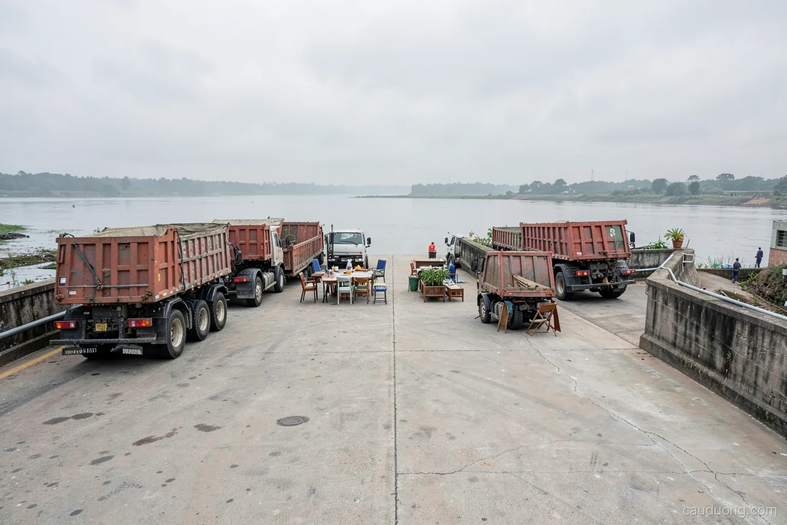 Several loaded dump trucks parked in a precise pattern on a concrete river bridge deck under overcast sky, engineers small in the distance, wide view from the riverbank